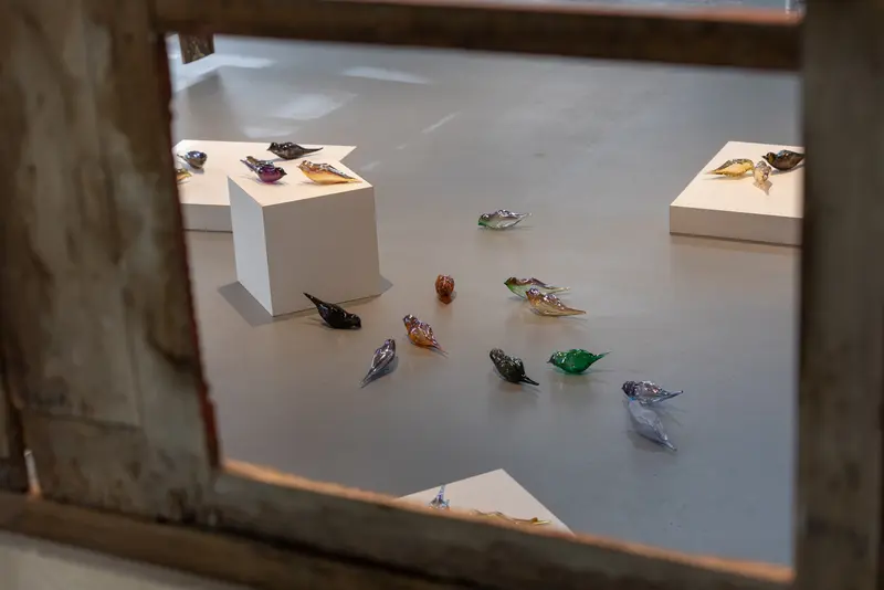 Blick durch ein Gitter auf eine Kunstinstallation mit gläsernen Vögeln